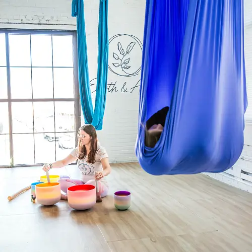 Woman resting in aerial yoga hammock with another woman playing sound bowls and doing a sound bath in the background