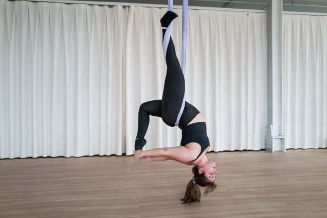 Aerial yoga student hanging upside down in a backbend pose with one leg hooked in the silk, inside a bright studio with white curtains.