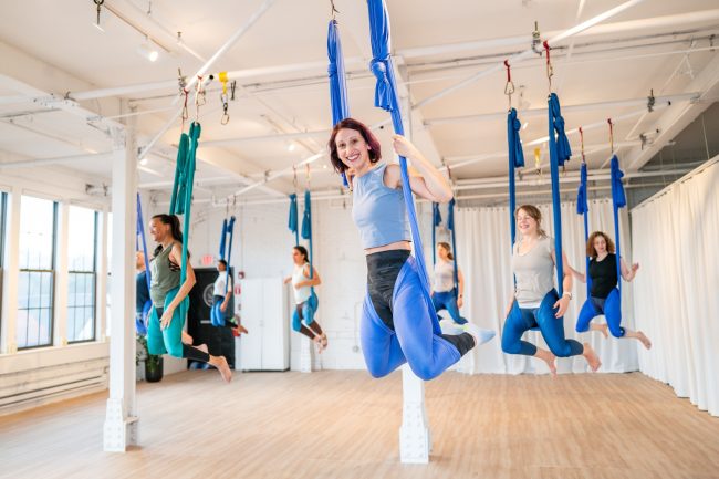 Group of students mid-swing in an aerial yoga class, smiling and suspended in blue silks during a joyful, energetic moment.